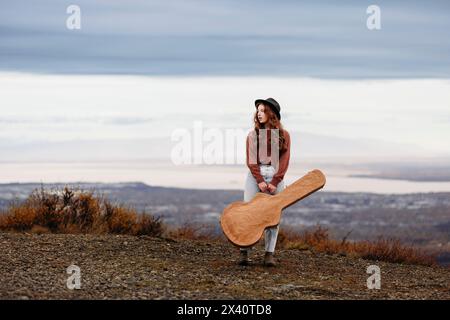 Portrait of a teenage girl wearing a black hat and blue jeans standing on mountain top holding her guitar case in the fall at Glen Alps Stock Photo