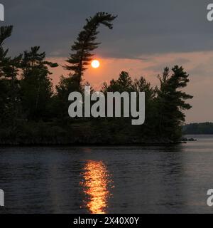 Setting sun reflects a warm sunbeam on the surface of lake water with silhouetted trees along the shoreline; Lake of the Woods, Ontario, Canada Stock Photo