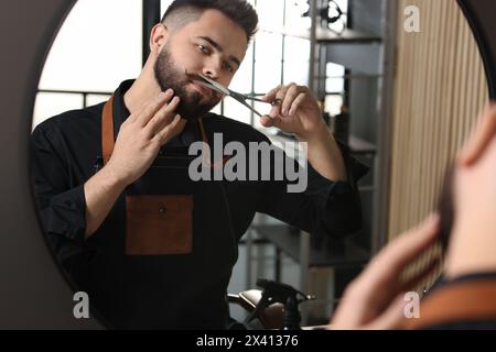Handsome young man trimming mustache with scissors near mirror indoors Stock Photo