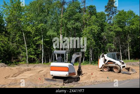 A compact front loader and mini excavator sits on the dirt at a new home construction lot, with trees in back of site, on a sunny day with blue sky. Stock Photo
