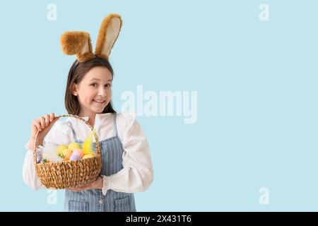 Happy little girl in bunny ears holding basket with Easter eggs on blue background Stock Photo