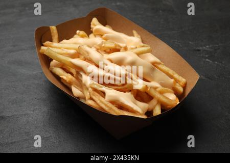 Tasty potato fries and cheese sauce in paper container on black table, closeup Stock Photo