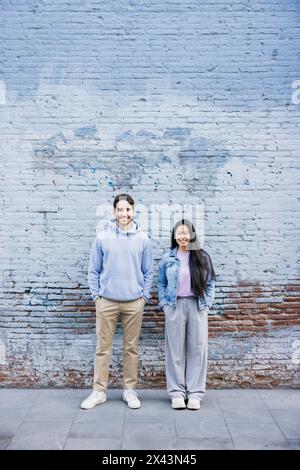 Young man and asian woman standing by blue brick wall. Couple with hands in pockets against a colored wall and smiling. Stock Photo