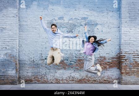 Young man and woman jumping by blue brick wall. Couple cheerfully jumping against a colored wall and smiling. Stock Photo