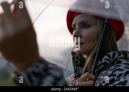 A woman wearing a red hat and a leopard print coat is holding a clear umbrella. The umbrella is open, and the woman is looking up at the sky. The Stock Photo