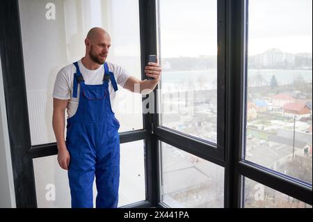 Portrait of young bald male construction worker in blue overalls using smartphone. Man standing near big window in apartment under renovation. Stock Photo