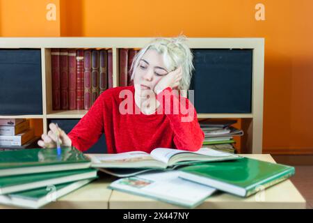 Young woman with blonde hair, looking tired, holding a pen while reading a book in a library setting, surrounded by books and showing signs of fatigue Stock Photo