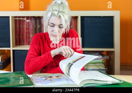 Young woman with bleached blonde hair, focusing on reading. Dressed in red, sitting at a library desk, surrounded by books, symbolizing deep concentra Stock Photo