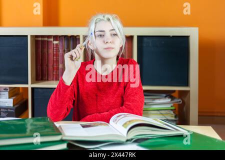 Thoughtful young woman with dyed blonde hair in a red sweater, contemplating while studying in a library, pen in hand, surrounded by books Stock Photo
