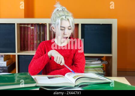 Young woman with blonde hair, wearing a red sweater, showing a surprised expression while pointing at a book in a library filled with shelves of books Stock Photo