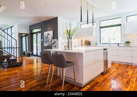 White high-gloss lacquered cabinets and island with grey leather bar stools and white quartz countertops in kitchen with acacia wood floorboards Stock Photo
