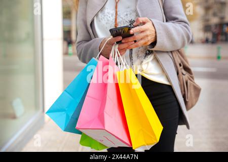 unrecognizable woman with shopping bags using an app on her smartphone outdoors. Stock Photo