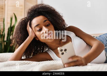 A curly African American woman in a tank top relaxing on a bed, holding a cellphone in a modern bedroom. Stock Photo