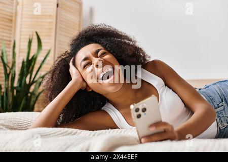Curly African American woman in a tank top relaxing on a bed, holding a smartphone in a modern bedroom. Stock Photo