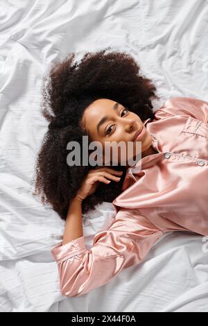 A curly African American woman in pajamas peacefully lays on top of a white sheet in her bedroom during morning time. Stock Photo