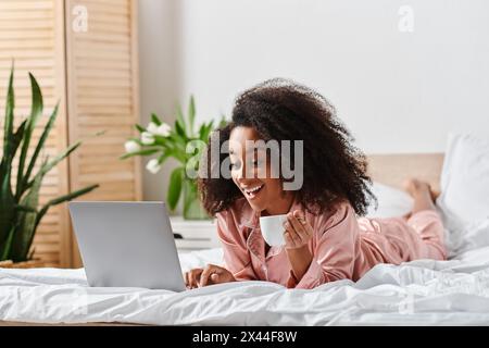 Curly African American woman in pajamas peacefully laying on bed, working on laptop in cozy bedroom during morning time. Stock Photo