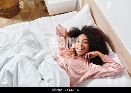 Curly African American woman in pajamas peacefully resting on a white bed in a serene bedroom during the morning. Stock Photo