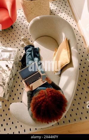 Directly above view of young woman with dyed red curly hair using laptop and smart phone in living room Stock Photo