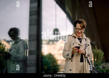 Smiling senior woman using smart phone while standing by glass reflection Stock Photo