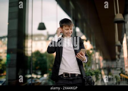 Smiling young businesswoman using smart phone while walking against building Stock Photo