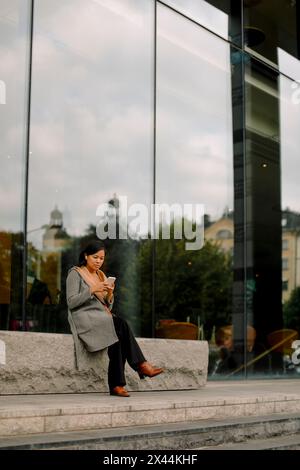 Full length of businesswoman using smart phone while sitting on bench Stock Photo