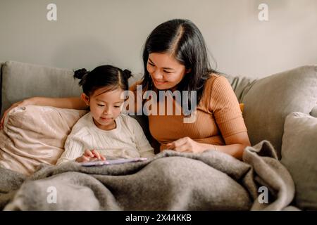 Smiling woman by daughter using digital tablet while sitting together on sofa at home Stock Photo