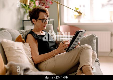 Retired senior woman using digital tablet while sitting on sofa at home Stock Photo
