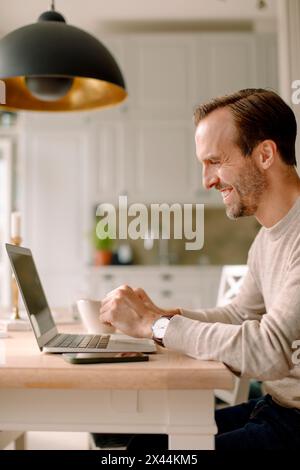 Side view of smiling businessman using laptop in kitchen while working at home Stock Photo