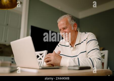 Smiling retired senior man using laptop at dining table in kitchen Stock Photo