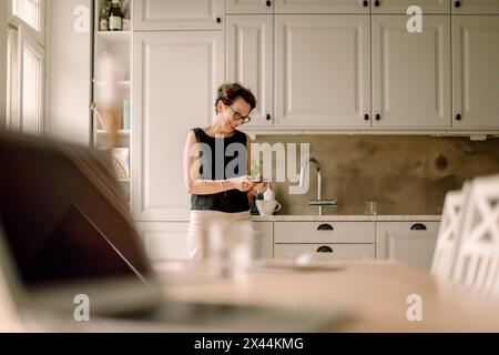 Retired senior woman using smart phone while leaning on kitchen counter Stock Photo