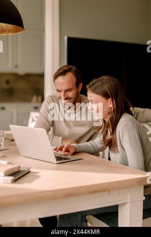 Smiling father sitting by daughter using laptop for studying at home Stock Photo