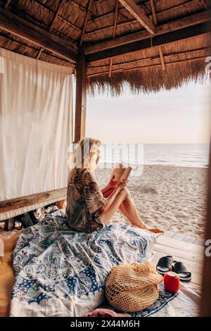 Woman reading book while sitting in cabana on vacation at beach Stock Photo