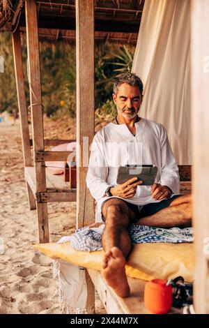 Mature man surfing net on tablet PC while sitting in wooden cabana on vacation at beach Stock Photo