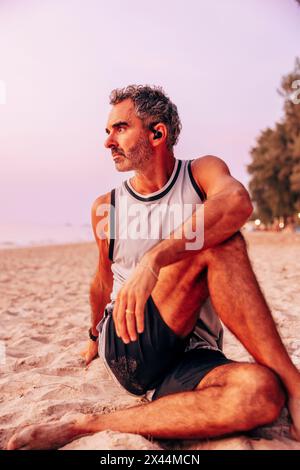 Mature man doing stretching while sitting on sand at beach Stock Photo
