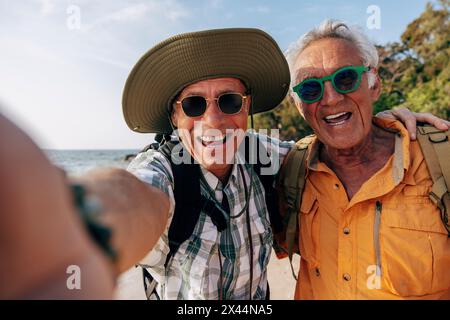 Portrait of happy senior gay couple wearing sunglasses and taking selfie at beach Stock Photo