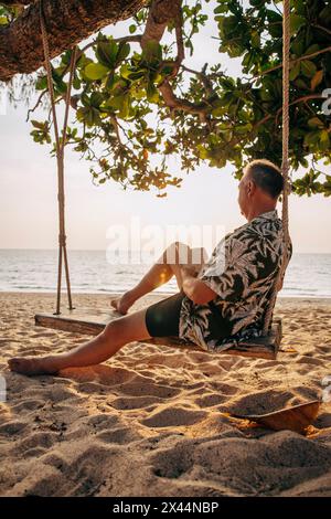 Side view of senior man reading book while sitting on swing at beach Stock Photo