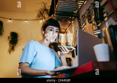 Female freelancer talking on speaker phone and using laptop while working at home office Stock Photo