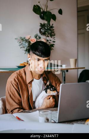 Smiling freelancer sitting with dog and working on laptop at home office Stock Photo