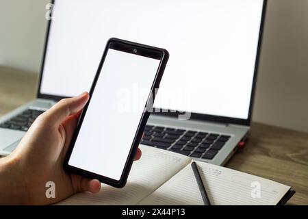 blank white screens of mobile phone and laptop, man holding phone in front of laptop at the table, scrolling , notebook and pen nearby Stock Photo
