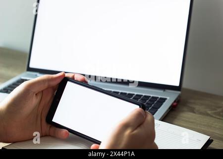 blank white screens of mobile phone and laptop, man holding phone in front of laptop at the table, scrolling , notebook and pen nearby Stock Photo