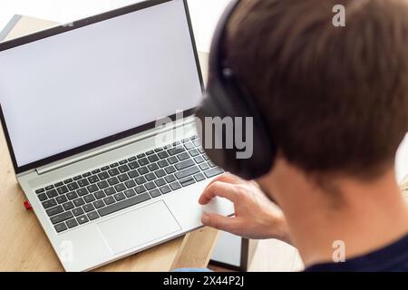 man wireless headphones sits in front of  blank white screen, at a small table, makes notes in notebook, image in light colors, online lesson or onlin Stock Photo
