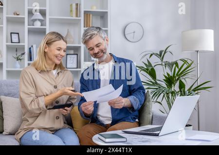 A mature couple in a cozy living room sorts and discusses documents with a laptop and calculator. They show teamwork and contentment while managing their finances. Stock Photo