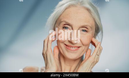 Medium Portrait: Senior Woman Looking at Camera, Touching, Massaging Beautiful Face, Smiling. Elderly Female with Grey Hair, Blue Eyes, Soft Skin. Dignified Old Age Concept for Natural Skin care Stock Photo