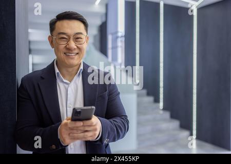 Confident Asian businessman in a suit smiles warmly while using his smartphone in the sleek, contemporary offices with stylish interiors. Stock Photo