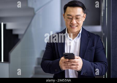 A cheerful Asian businessman in a dark suit is using his smartphone, standing in a stylish modern office setting, looking engaged and happy. Stock Photo