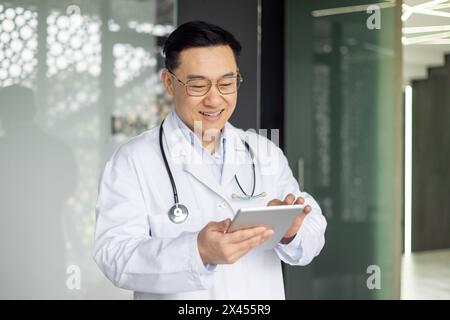 A cheerful Asian male doctor happily interacting with a digital tablet in the bright corridor of a contemporary hospital. Emphasizing professionalism and modern healthcare. Stock Photo