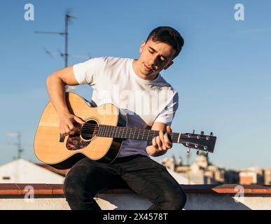 young man sitting playing acoustic guitar on a rooftop Stock Photo