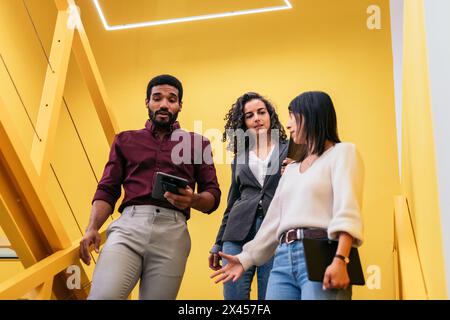 Multiracial coworkers happily talking about the company while walking down some yellow stairs in the office corridor Stock Photo