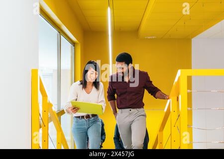 Multiracial coworkers happily talking about the company while walking down some yellow stairs in the office corridor Stock Photo