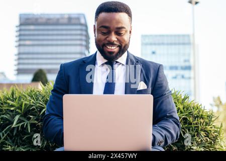 Front view of a smiling business black man in suit working with laptop outside Stock Photo